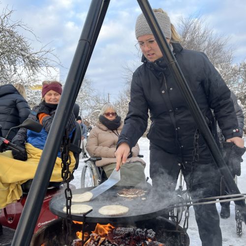 A staff member in a winter coat and hat is flipping Norwegian batter-based cakes on the outdoor fireplace. Three patients—one in a scooter, one in a wheelchair, and one standing—are in the background wearing winter coats and waiting to taste the delicious cakes.