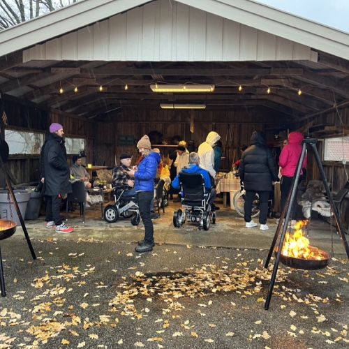 Two suspended, lit fireplaces flank an open wooden garage where several people in winter coats are gathered. Some people are standing, while others are in wheelchairs. Scattered autumn leaves cover the pavement in front of the garage.