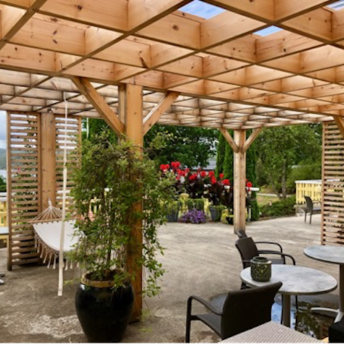 A large wooden pergola covers a central gathering place with several chairs, small tables, and a hammock. There are potted plants beneath the pergola and a view of flowers, trees, and water in the distance.