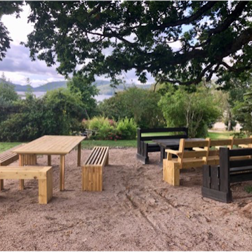 An outdoor seating area under a tree with light and dark brown wooden furniture, including two tables and several benches. The furniture is surrounded by verdant trees and grass. A cloudy sky is visible in the distance.