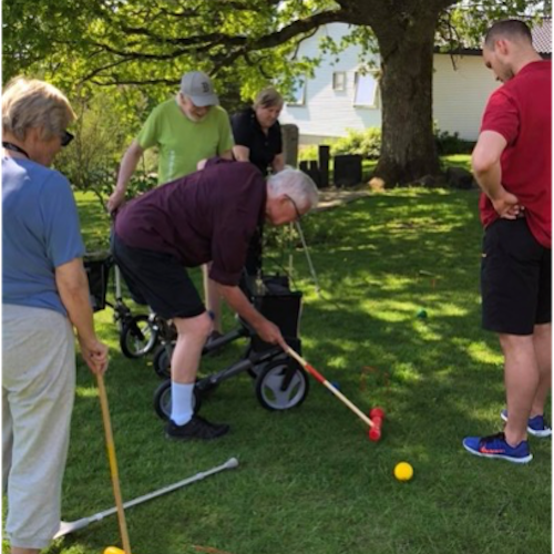 A group of people in summer clothes are standing with nearby ambulatory aids such as crutches, wheelchairs, and walkers. Two people are holding croquet mallets, with one leaning over and preparing to hit the yellow ball.