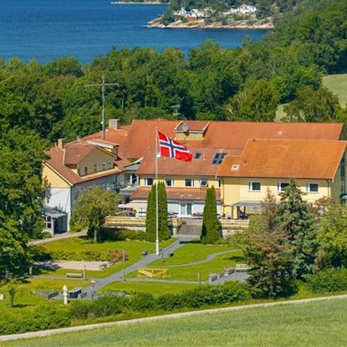 A photograph of the multi-story Unicare Bakke rehabilitation centre, with a garden featuring paths and a Norwegian flag in the foreground. Leafy trees and water are visible behind the building.