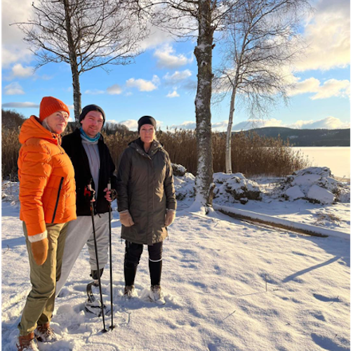 Three people wearing coats stand in a snowy winter landscape looking at the camera. The person in the middle has a prosthetic leg and is holding trekking poles. A few trees, hills, and water are visible in the background.