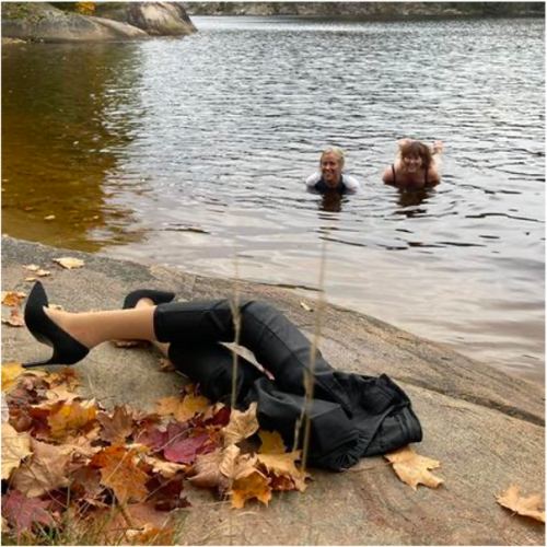  Two people are floating in the water and smiling at the camera. In the foreground, there is a pair of prosthetic legs wearing black pants and black high heels on the smooth rock shore amidst scattered autumn leaves.