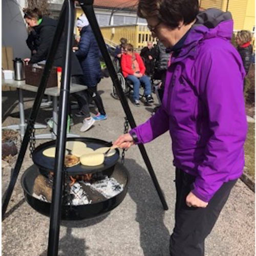 A person in a purple jacket is flipping batter on a suspended fireplace outside. In the background, there is a coffee carafe on a table and a group of people in winter clothes standing or in wheelchairs.