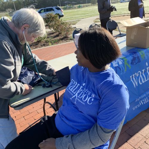 A woman in a blue Walk with a Doc t-shirt sits at an outdoor table where she is having her blood pressure tested. The table has a Walk with a Doc banner, and bottles of water lined up in a row.