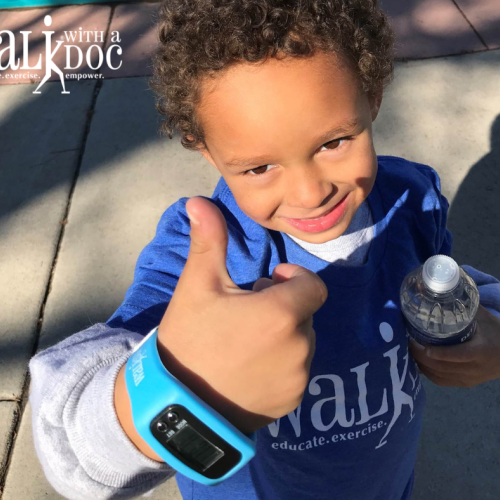 A child smiles up at the camera giving a thumbs up. The child is wearing a Walk with a Doc t-shirt and holding a bottle of water.