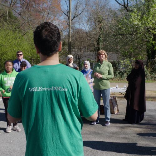 A Walk with a Doc leader with their back to the camera addresses a group of people of varying ages and ethnicities, standing around him in a semi-circle.