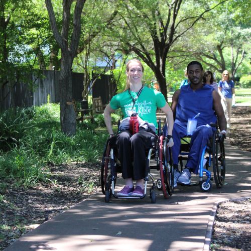 Two wheelchair users face the camera on a footpath surrounded by trees and shrubs. Behind them, several people are walking in the bright sunshine.