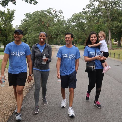 Four adults wearing blue Walk with a Doc t-shirts, one of whom is carrying a small child, are walking on a small road with trees on either side. In the background, there is a lake.