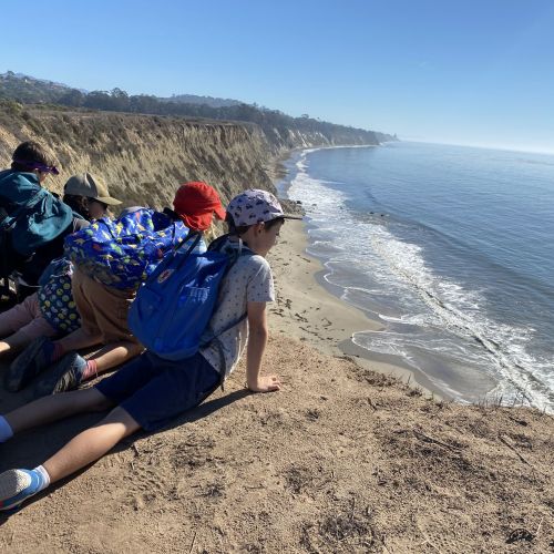 Four kids in hiking clothes lay by the edge of a cliff that overlooks the ocean. They eagerly admire the landscape spread before them, which includes a sunny, cloudless sky and a vividly blue ocean that disappears into the horizon.