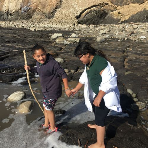 Two youths hold hands as they stand in a shallow lake. One of them holds a hiking stick while glancing back at their friend in reassurance. The lake itself has rocks of various sizes scattered throughout, and the lakeshore is composed of even more rocks.