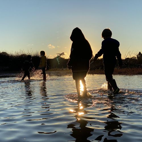 Four kids wade through a lake, which glimmers orange from the sunset reflecting across the surface. The sun, hidden behind a kid’s silhouette, sinks below a line of trees that border the lake, thus casting the entire landscape in shadows.