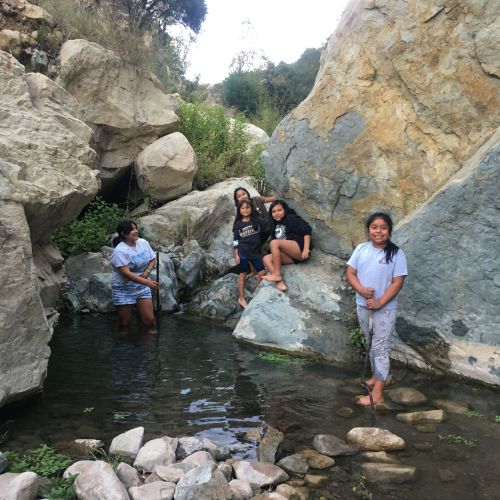 A group of youths take a break in a shady enclave formed by towering rocks. Three of them are standing up and leaning against the rocks while the fourth chooses to sit on a boulder and rest her legs. Grass peeks out from between the boulders that surround the group, and the ground is covered with water.