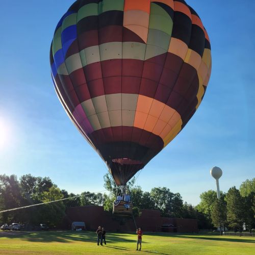 In an open green space, a colorful hot air balloon is suspended above three people, one of whom is holding a rope attached to the balloon’s basket.