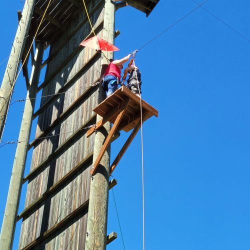 On a high ledge attached to a large, wooden climbing wall, two people stand attached to a zip line by multiple wires.