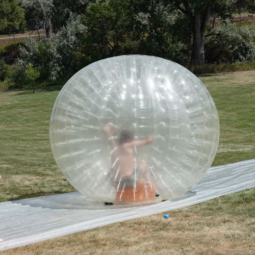 On a plastic slip n’ slide one young person stands and watches another slide along inside a zorb. In the background, an open grassy spaced is backed by trees and hedges.