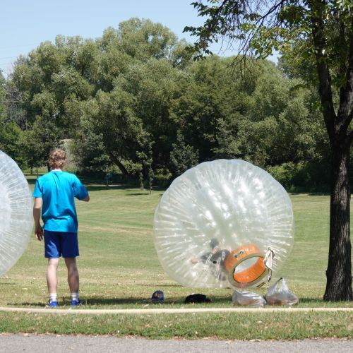 Two young people in shorts and t-shirts stand and are looking at two zorbs in front of a green lawn surrounded by trees.